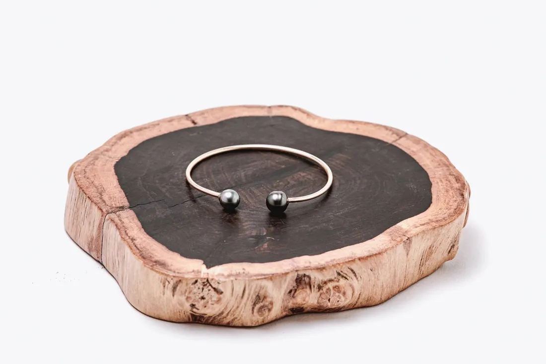 Mily open bangle with two black Tahitian pearls resting on a slab of koa wood